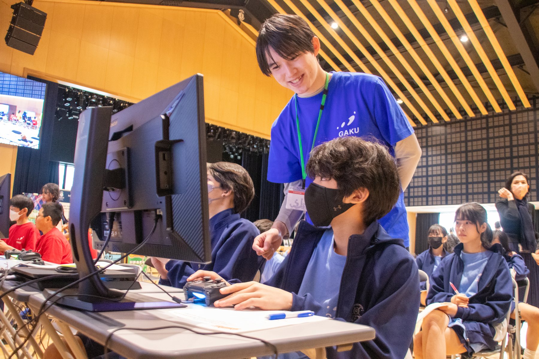 A GAKU coach in branded shirt assisting a student at a Puyo Puyo programming station